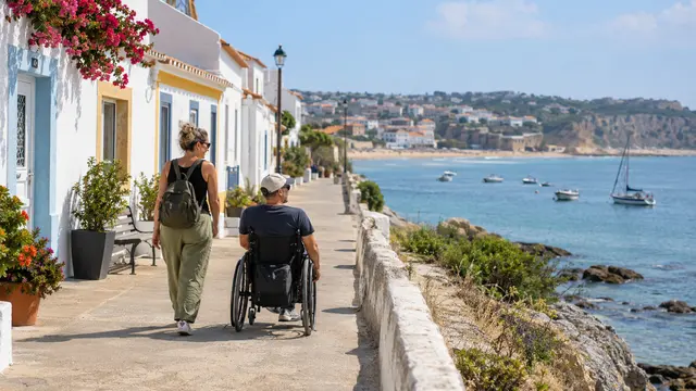 An adult wheelchair user with a companion following a smooth step-free promenade beside low Portuguese homes.