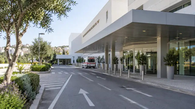 A clear road leading to a contemporary hospital entrance with a stationary unbranded ambulance in the far background.