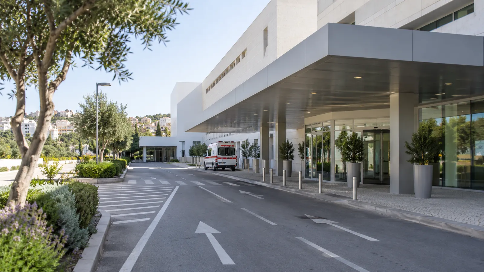 A clear road leading to a contemporary hospital entrance with a stationary unbranded ambulance in the far background.