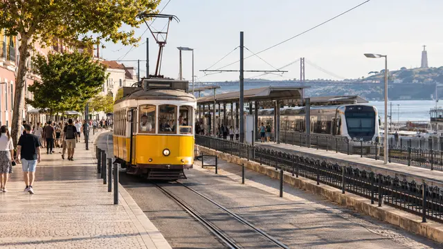 A Portuguese city tram passing a broad pedestrian pavement beside a suburban train connection.
